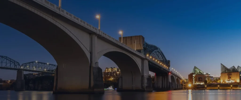 A concrete arch bridge over the Tennessee River in Chattanooga at dusk, with the lit riverfront beyond.